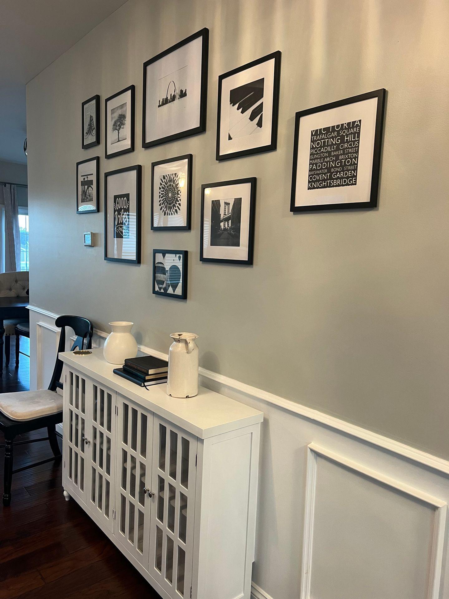 Wall with framed black and white artworks above a white cabinet with books and vases.