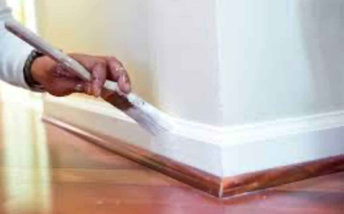 Person painting white baseboard trim with a brush on a wooden floor.