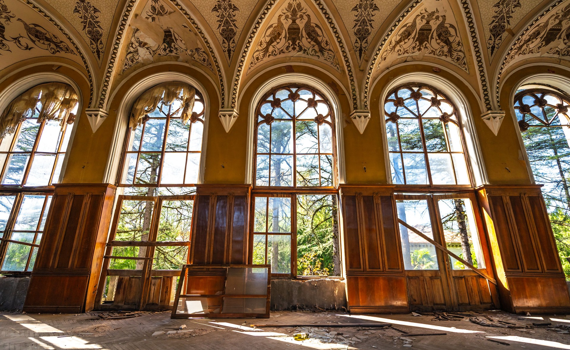 Abandoned historical hall with ornate vaulted ceiling, large arched windows, wooden paneling, and peeling paint, sunlight streaming through